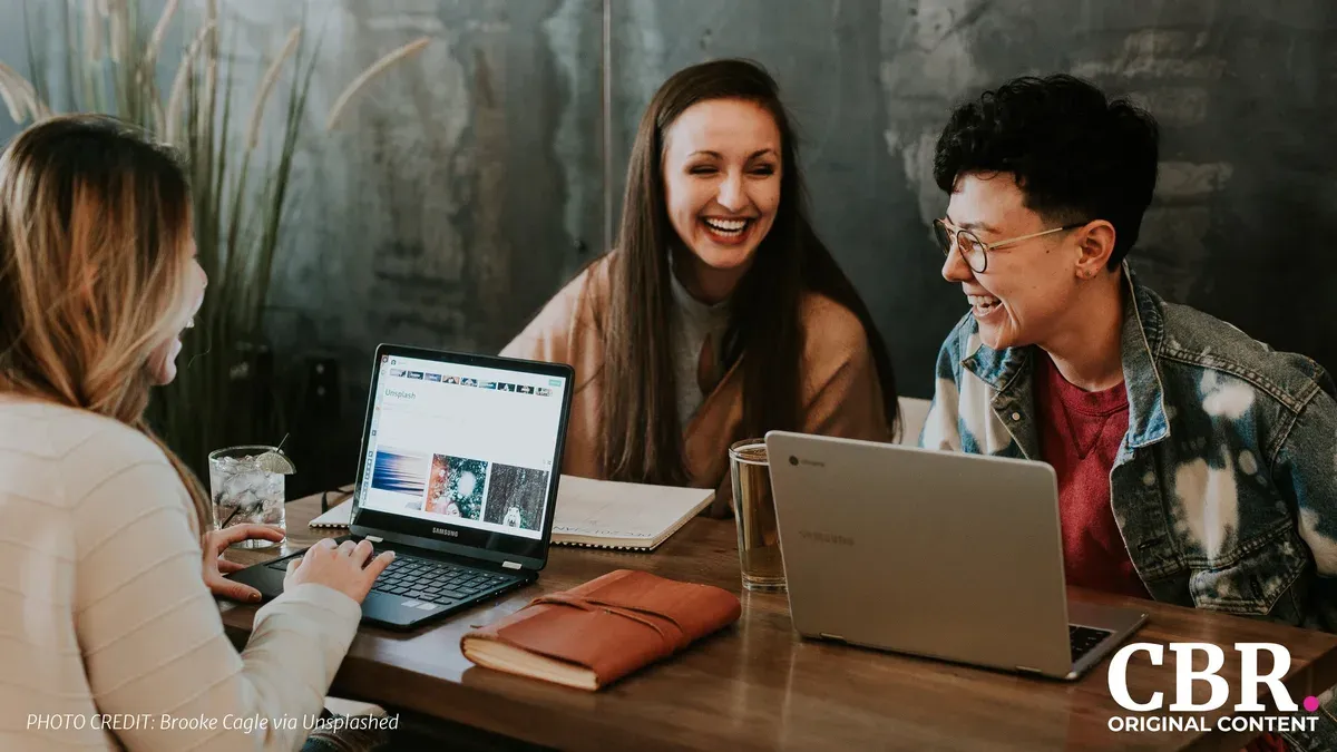 Three colleagues laughing together while looking at laptop screens at a table