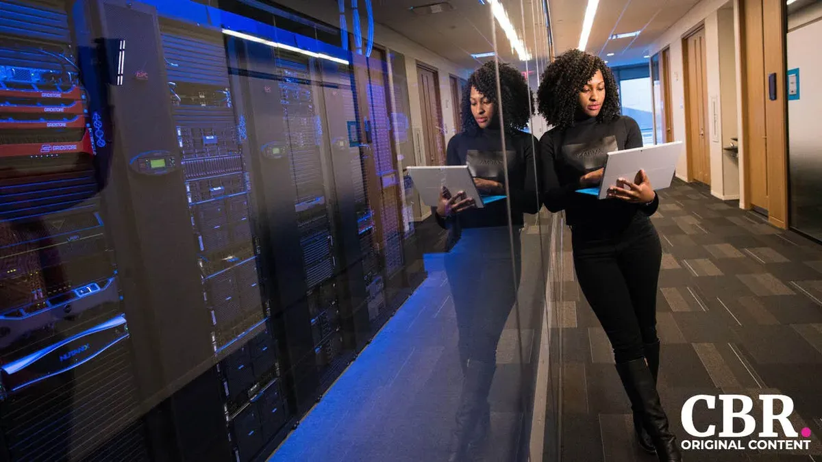 A woman reviewing a laptop in a data center corridor, reflected in the glass
