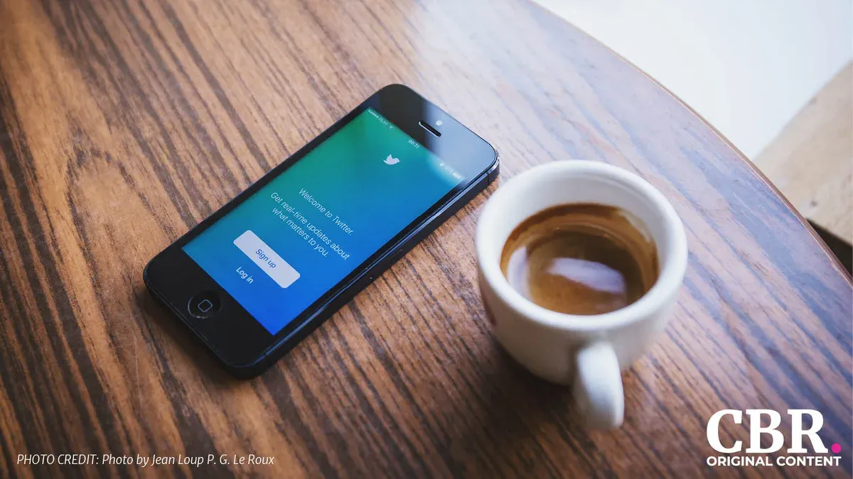 A smartphone showing the Twitter sign-up screen next to a cup of coffee on a wooden table